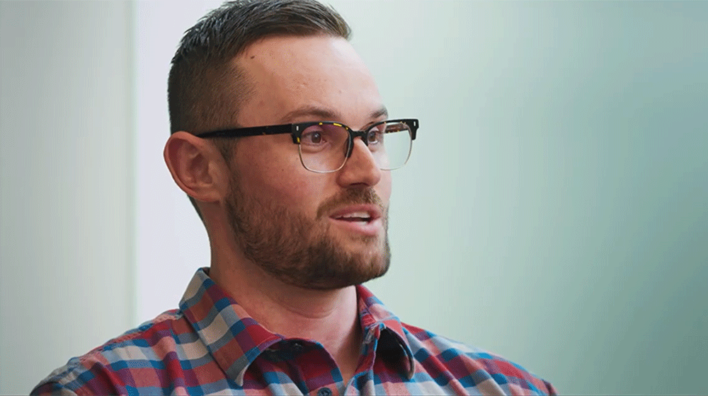 Side head shot of a man with short brown hair and glasses, wearing a plaid button-down shirt
