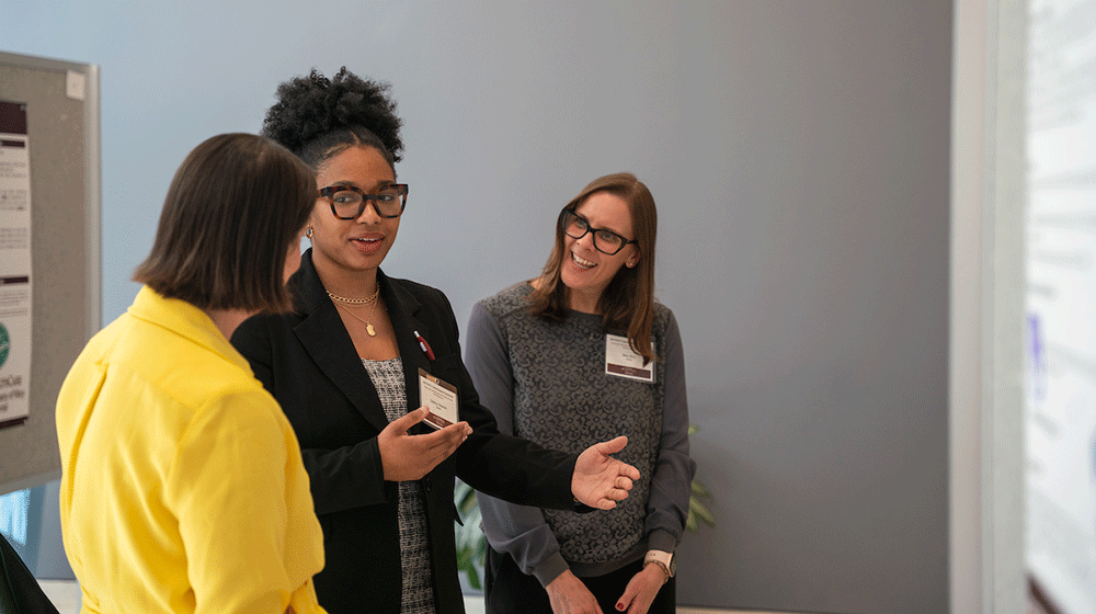 Three women stand and talk in front of a research poster display