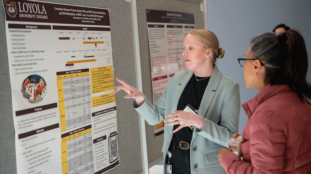 A student points at a research poster and explains it to a woman looking on