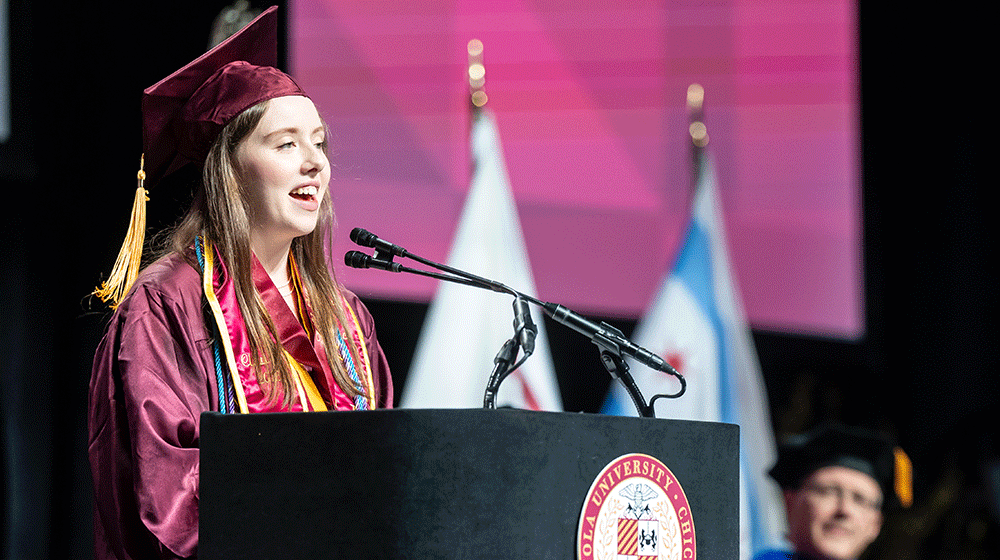 Kate Heneghan wears graduation robe and cap and stands at a podium speaking