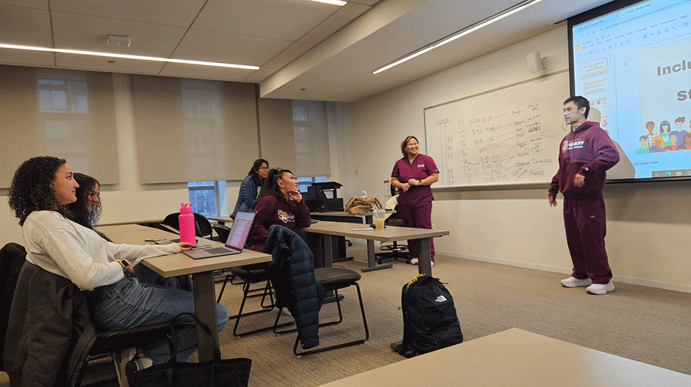 Students sit in a classroom and listen to a student in scrubs, standing, speak at the front of the classroom