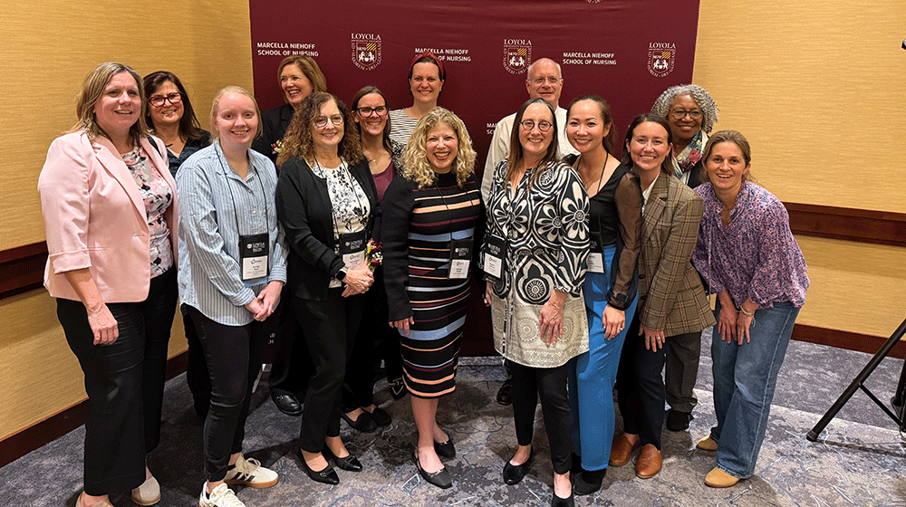 Faculty stand in front of a Loyola Nursing maroon backdrop and smile