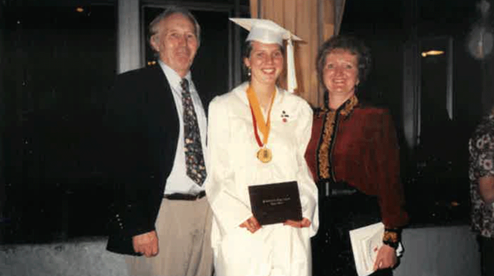Melissa Murphey stands in white graduation gown with her father standing to her right and her mother to her left.