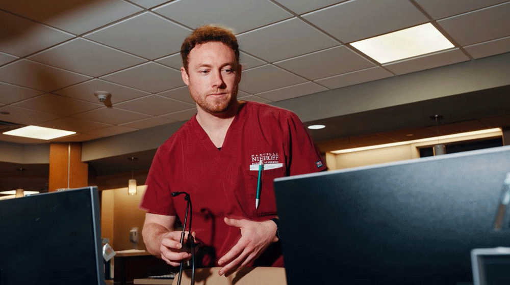 A male nurse in maroon scrubs holds a stethoscope and looks at a computer monitor in an office