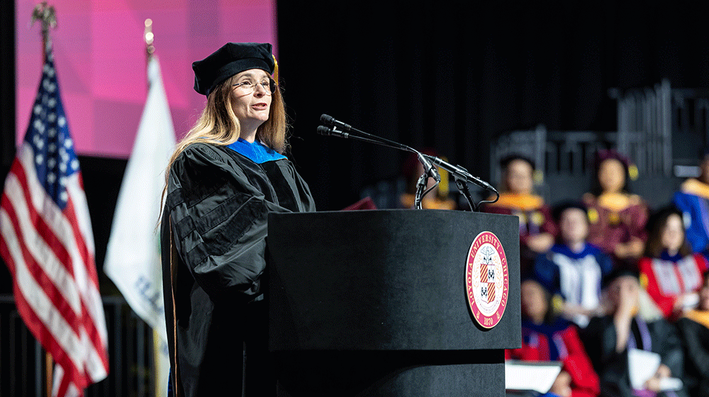 Lauren R. Source, wearing black cap and gown, stands at a podium and speaks during Commencement