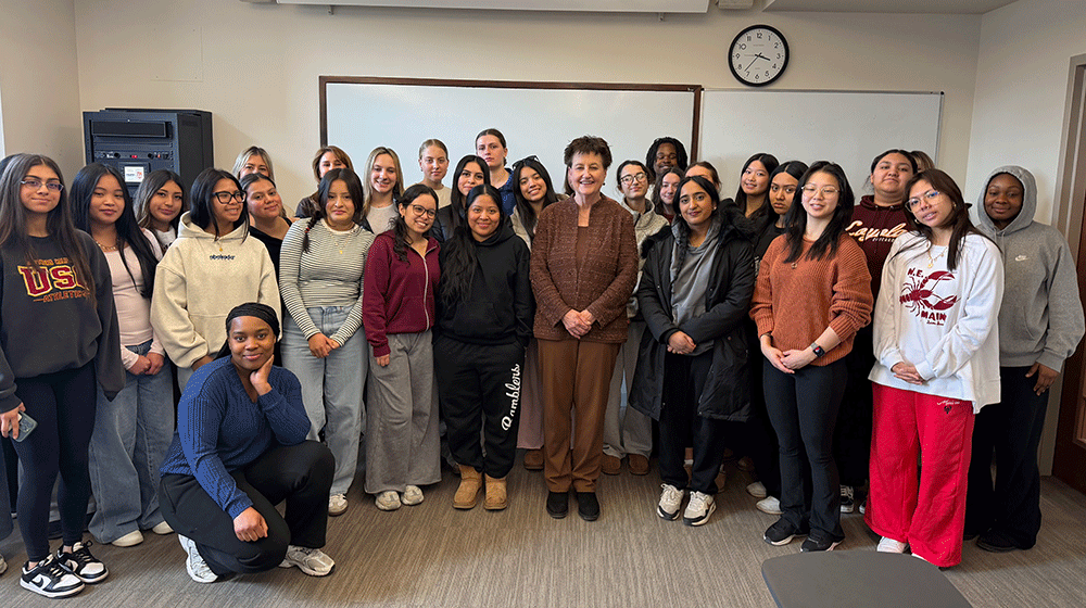 The dean stands in front of a white board in a classroom with college students, mostly female, beside her