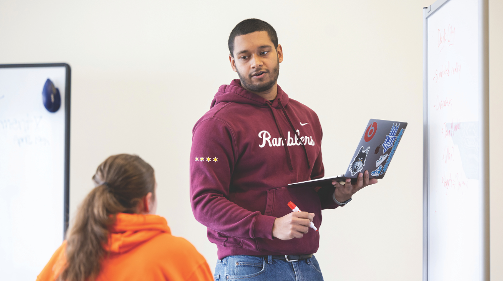 Tutor standing holding laptop and marker in front of a dry erase board turned to speak to sitting student