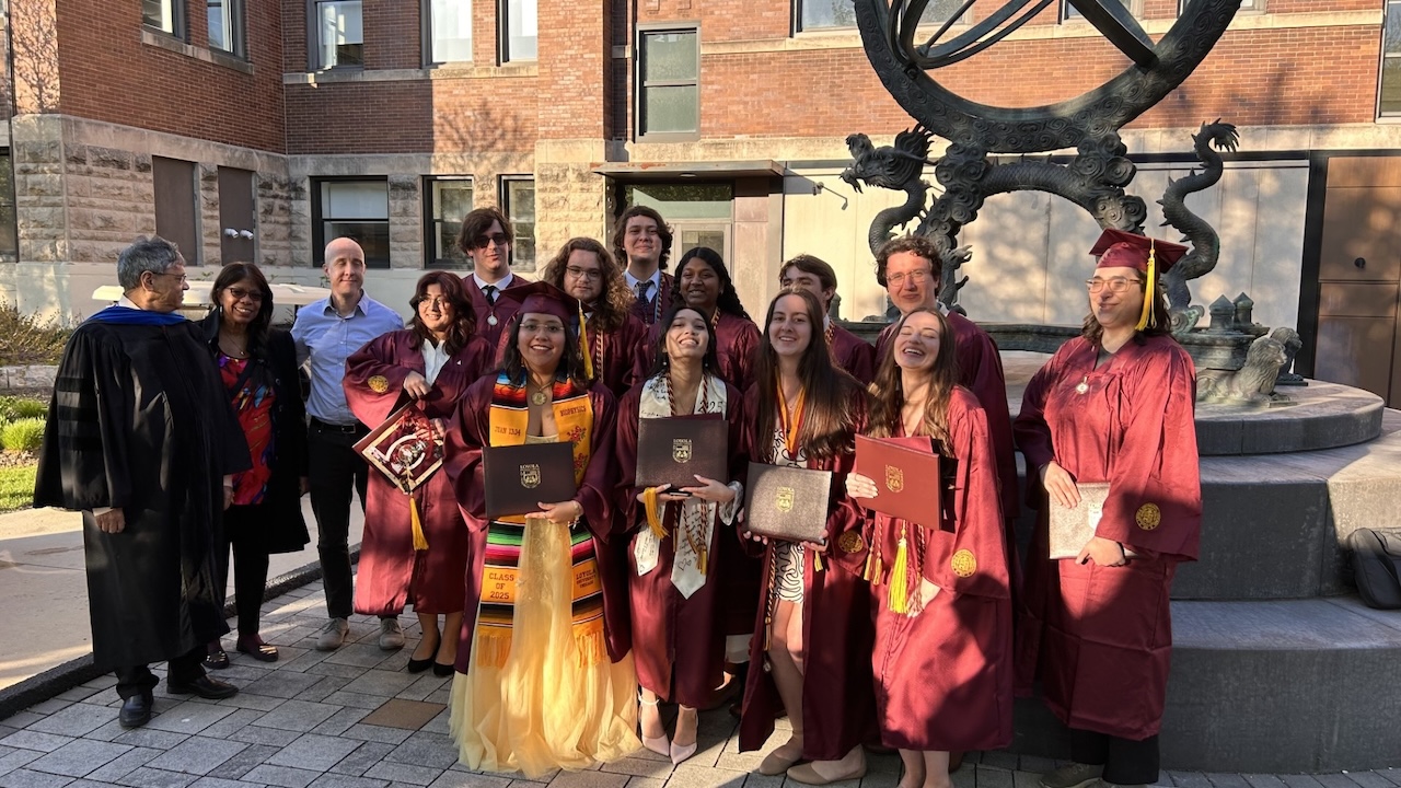 Group of graduates in maroon caps and gowns holding diplomas and smiling outdoors in front of a sculpture.