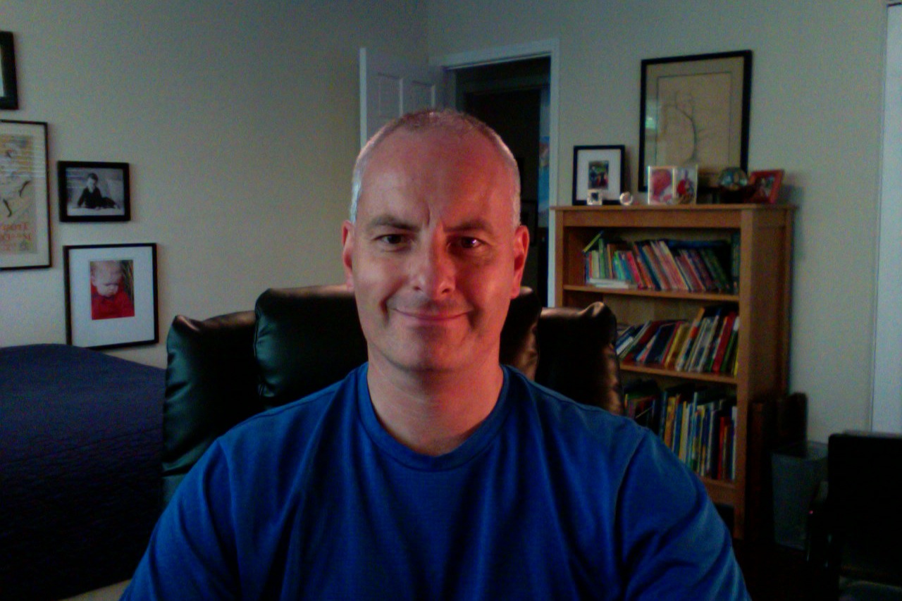 A man in a blue shirt sits in a chair at home, with a bookshelf, framed photos, and artwork in the background