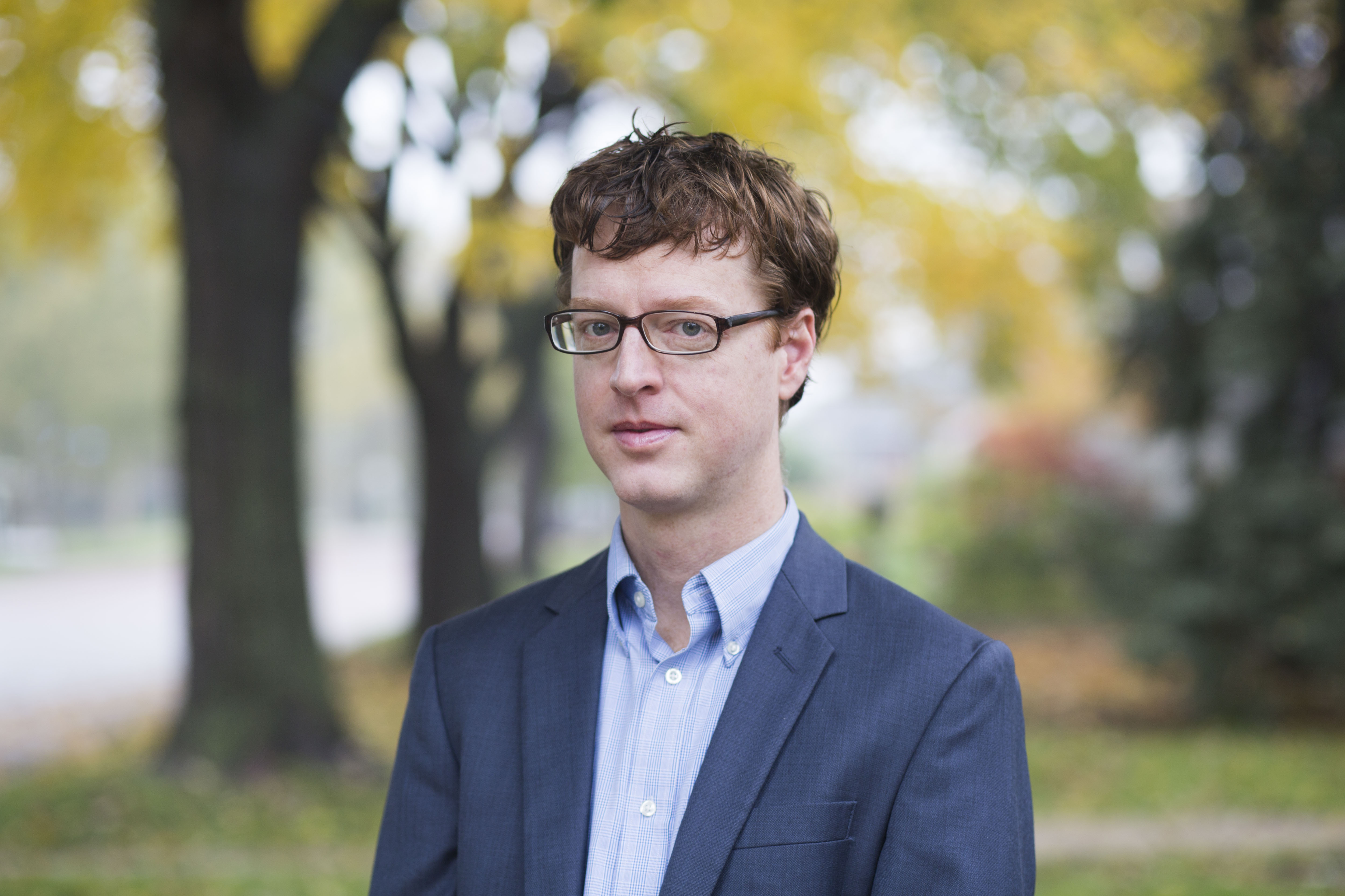 Man wearing glasses and a blazer standing outdoors with trees in background