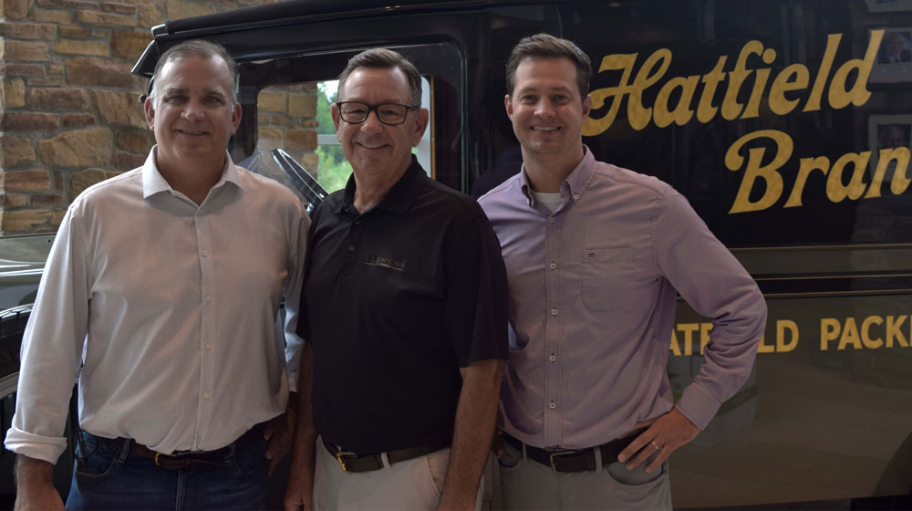 Three men in business attire pose for a photo in front of a truck and brick wall