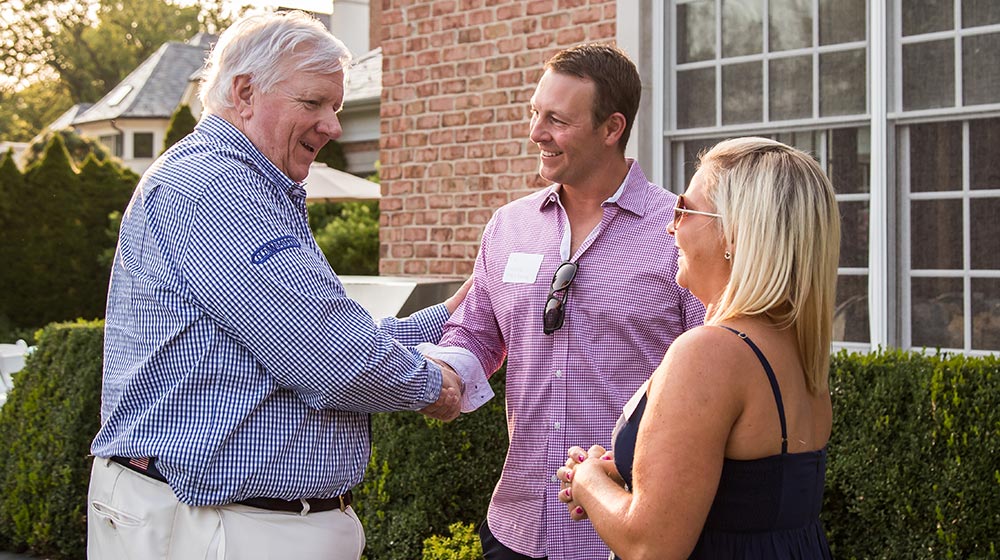 Three people are outdoors engaged in conversation. An older man is shaking hands with a younger man, both smiling. A woman stands nearby, also smiling. The background includes greenery, a brick building, and large windows.