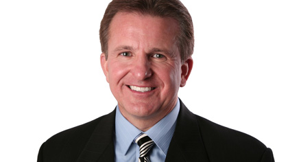 Headshot of a Quinlan faculty member smiling, wearing a dark suit jacket, light blue shirt, and striped tie against a light gray background.