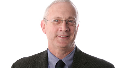 Headshot of a Quinlan faculty member wearing glasses, a dark blazer, light blue shirt, and dark tie, smiling against a light gray background.