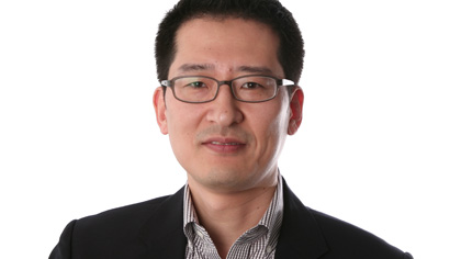 Headshot of a Quinlan faculty member wearing rectangular glasses and a dark blazer over a patterned shirt, looking at the camera against a light gray background.
