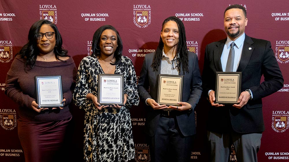 Four award winners smiling while holding their awards and standing in front of a Quinlan School of Business backdrop