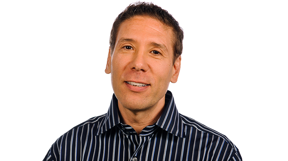Headshot of a Quinlan faculty member with short dark hair, wearing a dark striped button-down shirt, looking at the camera against a light gray background.
