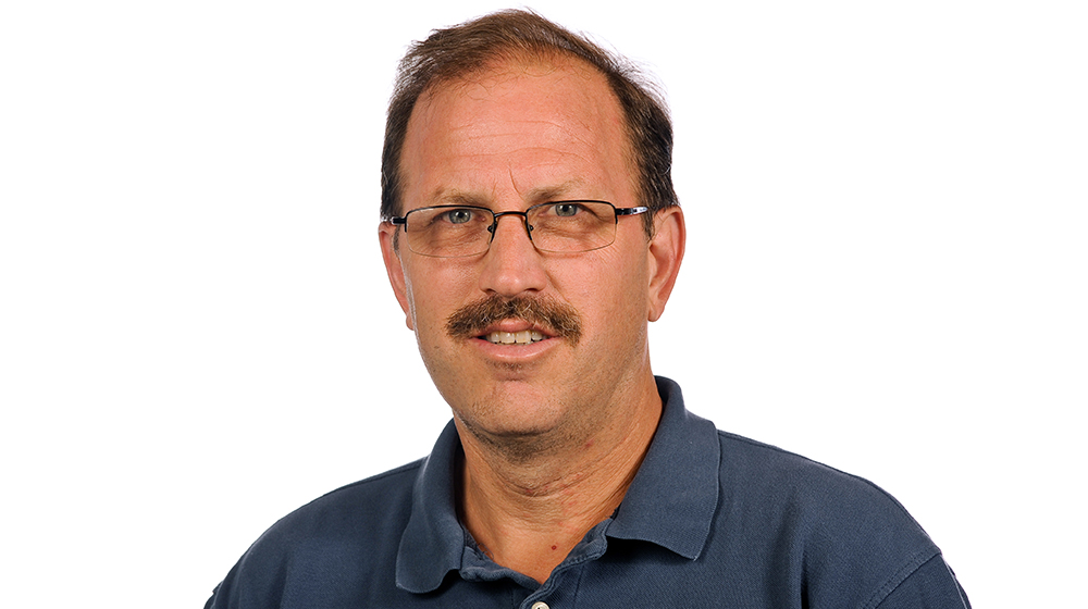 Headshot of a Quinlan faculty member with glasses and a mustache, wearing a dark blue polo shirt, looking at the camera against a light gray background.