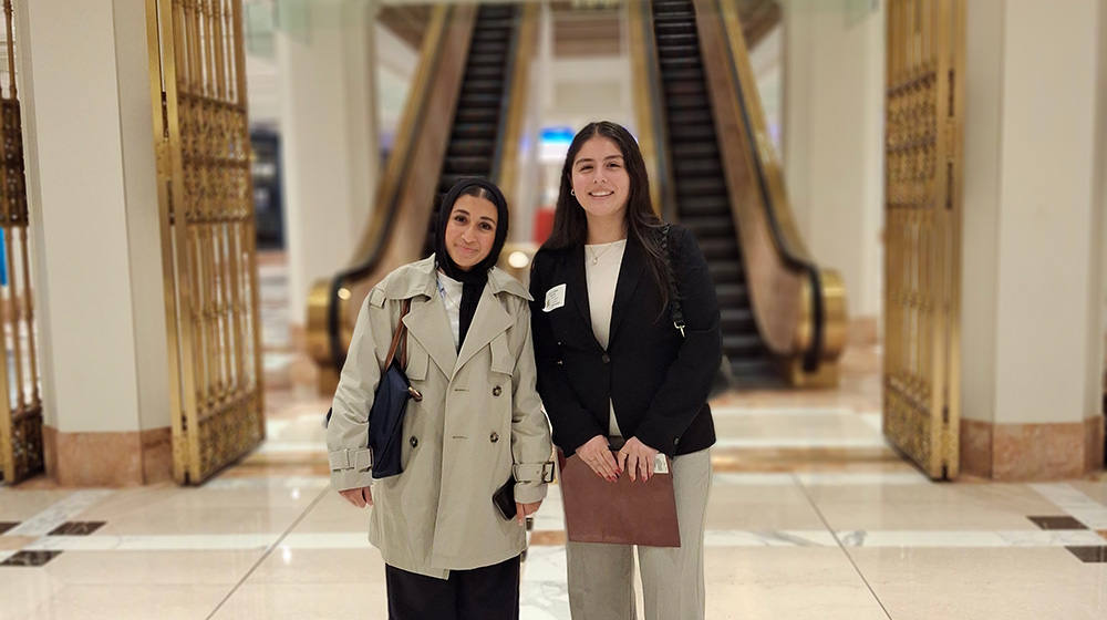 Two quinlan students, smiling and standing side by side, in the Wintrust building, home to Greenwood Project. The student to the left is wearing a tan overcoat, the student to the right is wearing a black blazer.