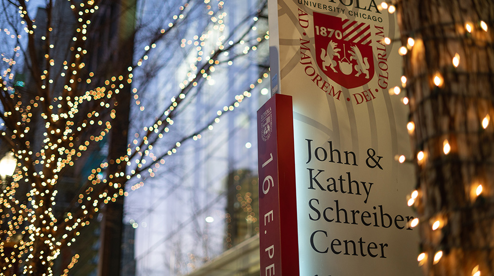 Sign for the John and Kathy Schreiber Center at dusk with christmas lights shining in the background.