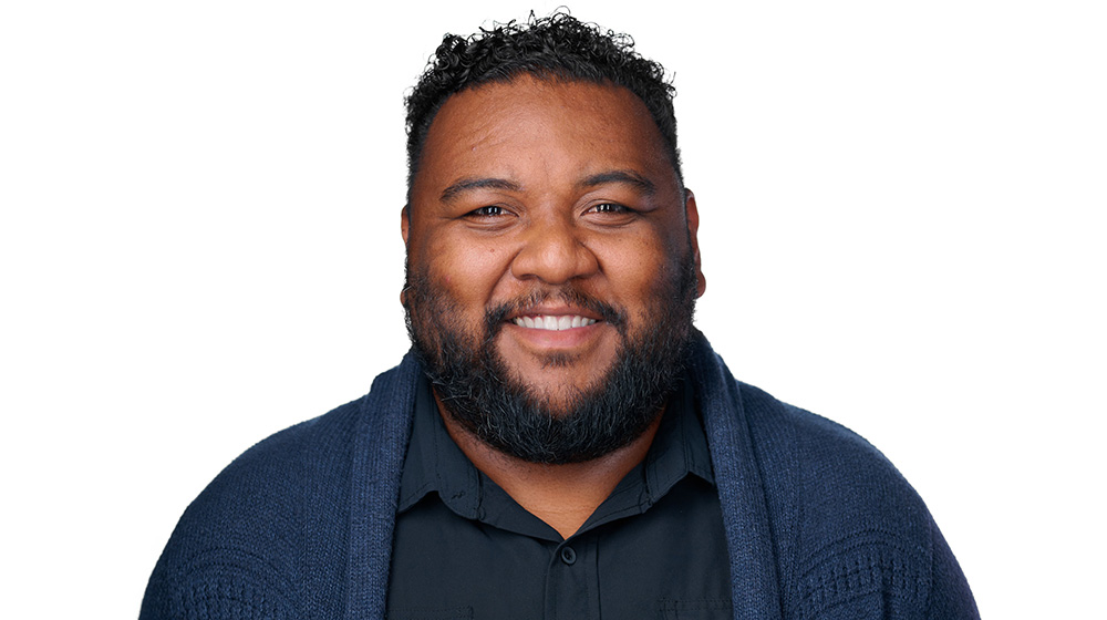 Headshot of a Quinlan faculty member with short curly hair and a beard, smiling, wearing a black shirt and dark cardigan against a light gray background.