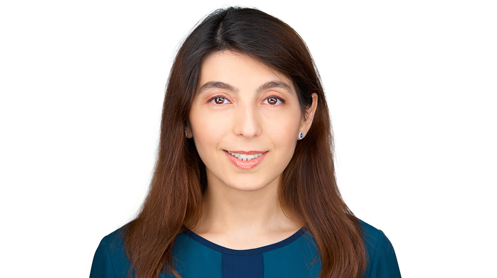 Headshot of a Quinlan faculty member with long brown hair, wearing a teal top and small stud earrings, smiling at the camera against a light gray background.