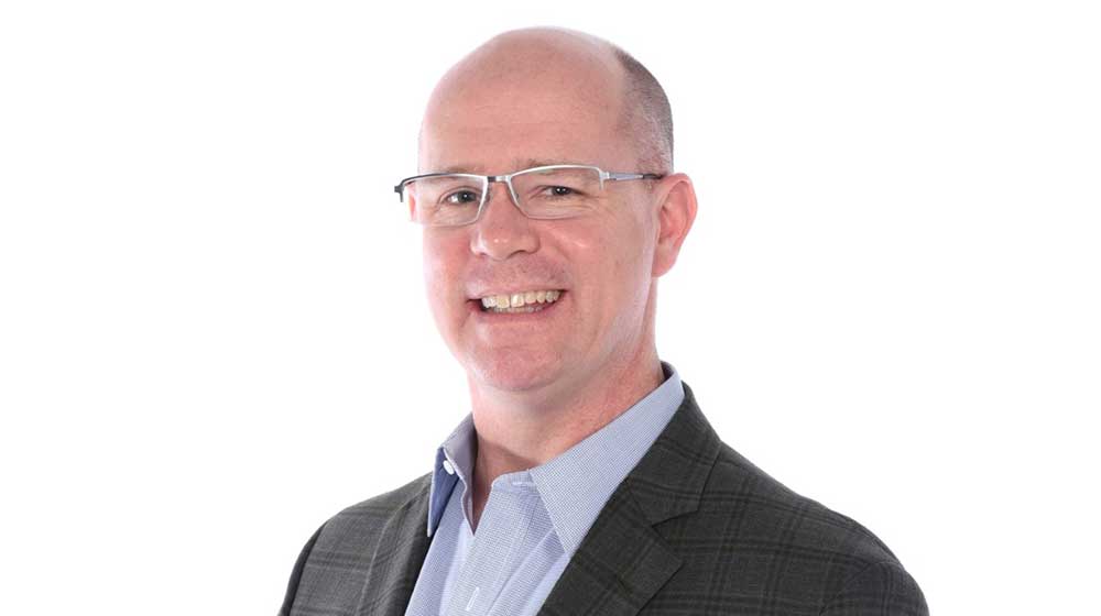 Headshot of a Quinlan faculty member with a shaved head, wearing glasses, a light blue shirt, and a dark plaid blazer, smiling against a light gray background.