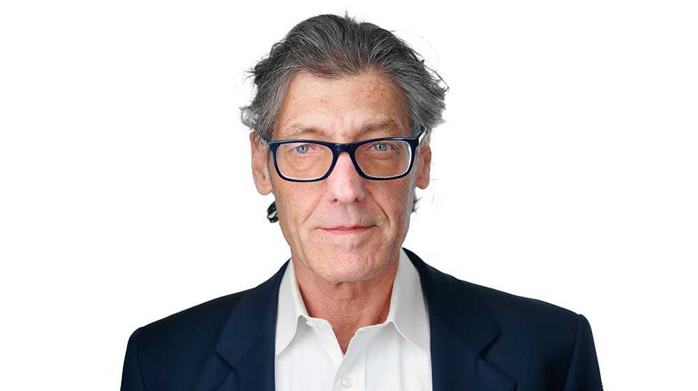 Headshot of a Quinlan faculty member with gray hair and blue-framed glasses, wearing a dark blazer over a white shirt, facing the camera against a light gray background.