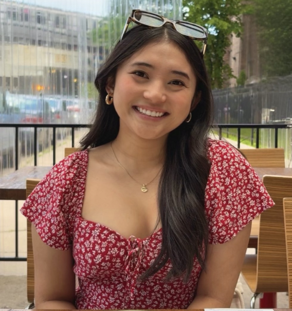 A female student is outside at a restaurant sitting at a table wearing a red floral shirt and sunglasses, smiling at the camera.