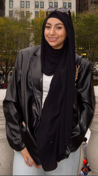 A female student is standing outside in downtown Chicago in a black jacket and blue jeans with sunglasses, smiling at the camera.