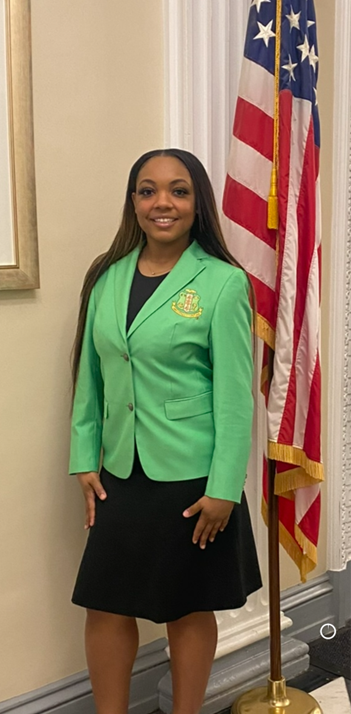 A female student is standing indoors next to an American flag and wearing a bright green blazer.