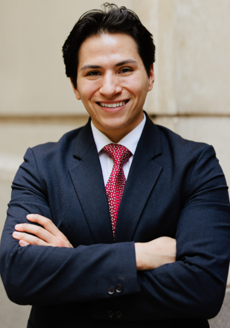 A male student is standing outside a building wearing a black blazer and red tie with his arms folded across his chest, smiling at the camera.