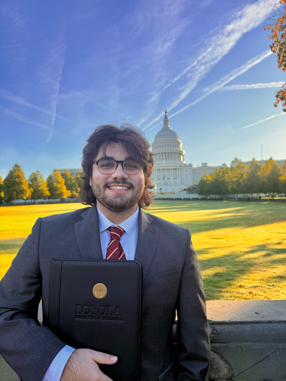 A male student is standing outside of the White House wearing a grey blazer and red tie while carrying a Loyola padfolio, smiling at the camera.