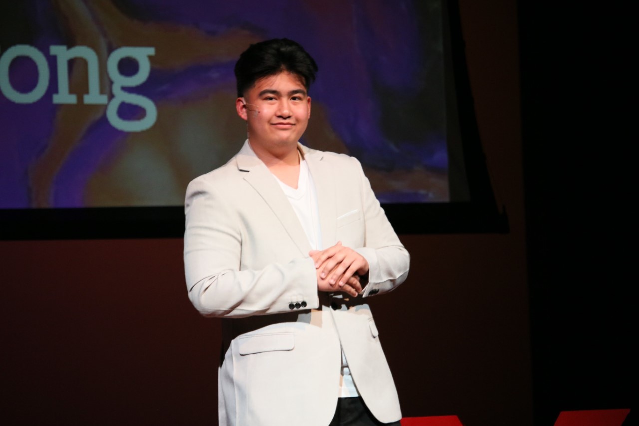 A male student is standing on a stage in a white jacket with his hands in front of his body and is smiling at the camera.