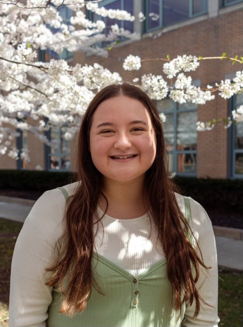 A female student is outside near a blossoming tree with flowers wearing a white and green shirt, smiling at the camera.