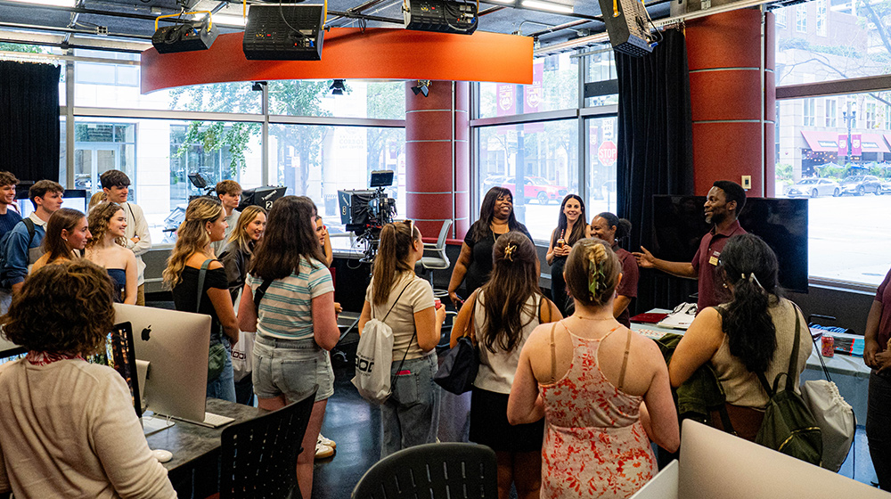 A group of students gathers inside a media studio as a guide speaks to them, surrounded by cameras, computers, and large front windows.