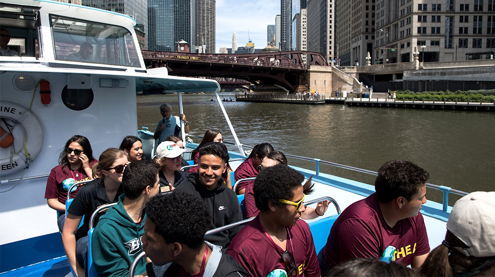 A group of people enjoy a boat tour along a city river, passing tall downtown buildings and a historic raised bridge.