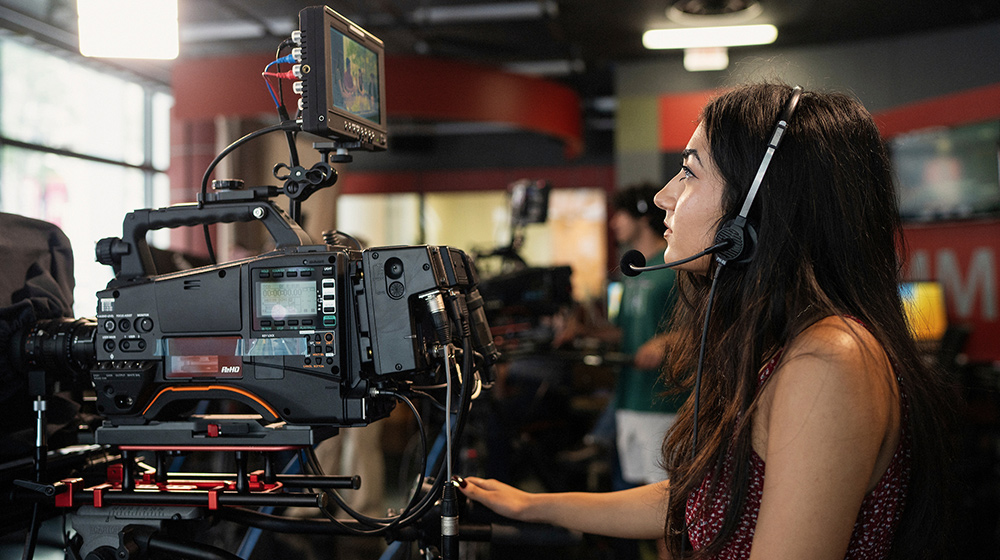 A person wearing a headset operates a professional video camera inside a studio filled with equipment and crew during filming.