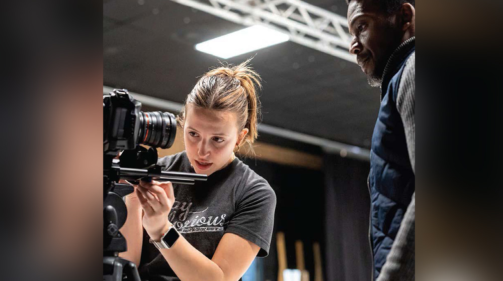 Person adjusting a professional video camera on a tripod in a studio setting while preparing equipment alongside another individual.