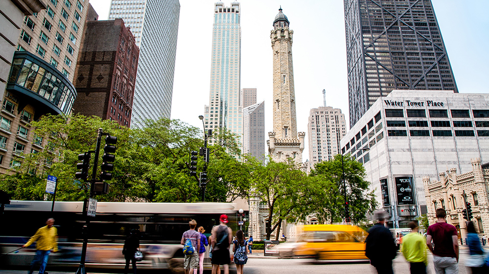 Busy city intersection with pedestrians, buses, and cars moving past Chicago’s historic Water Tower surrounded by tall modern skyscrapers.