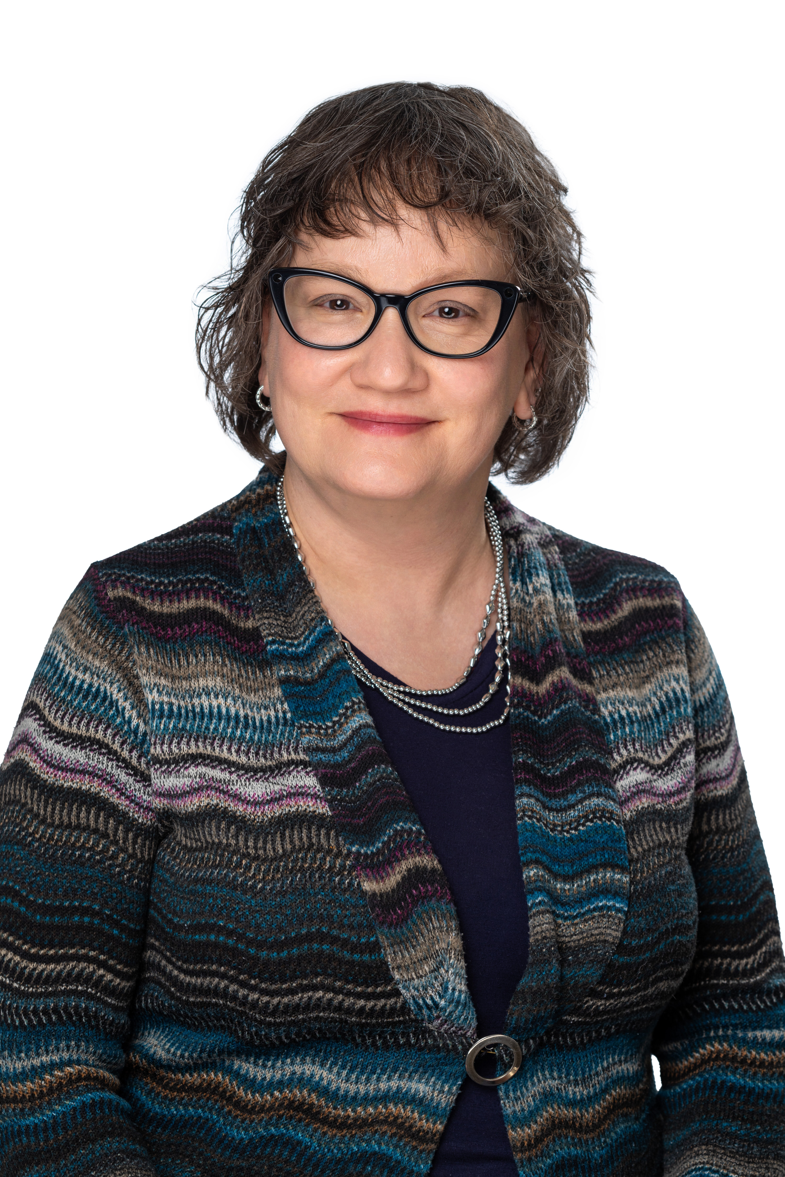 Headshot of a School of Social Work faculty member with short wavy brown hair and dark-framed glasses, wearing a multicolored patterned cardigan over a navy top and layered necklaces, smiling against a light gray background.