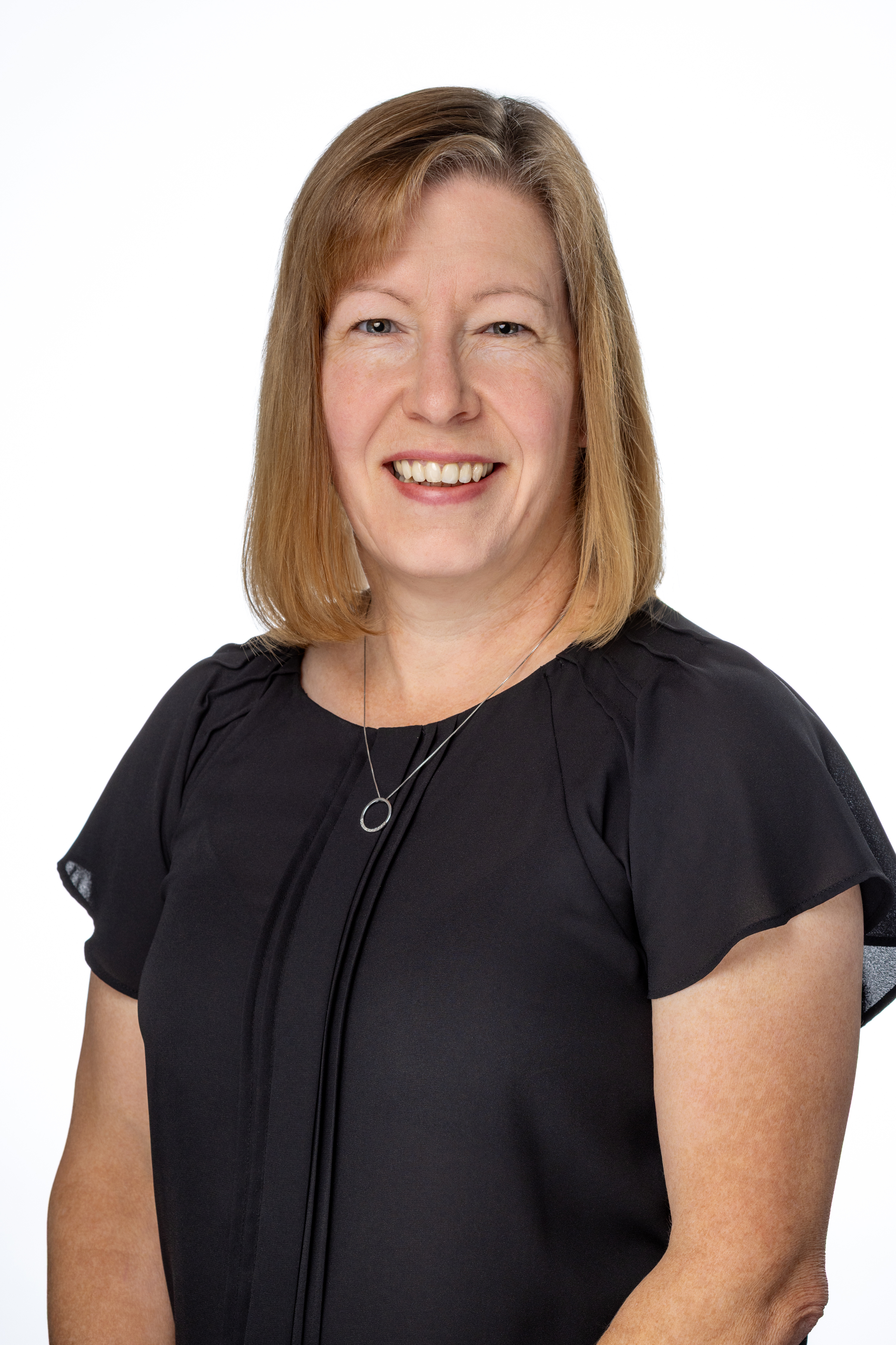 Christie Mason, a woman with shoulder-length light brown hair, wearing a black short-sleeve blouse and a simple circular pendant necklace, smiling against a light gray background.