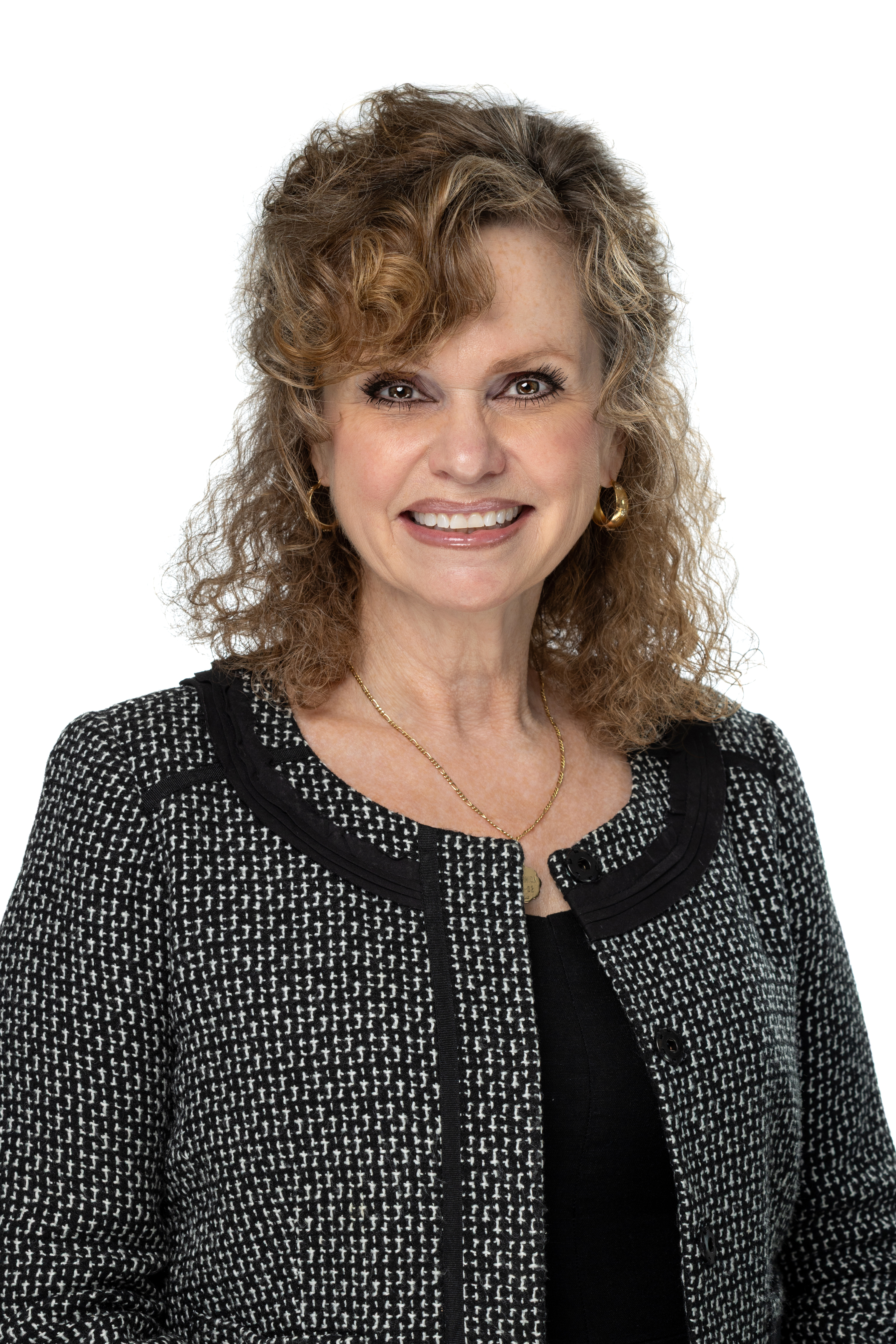 Maria Vidal de Haymes, PhD, smiling and wearing a black top with a textured black-and-white jacket, gold hoop earrings, and a delicate gold necklace, against a light gray background.