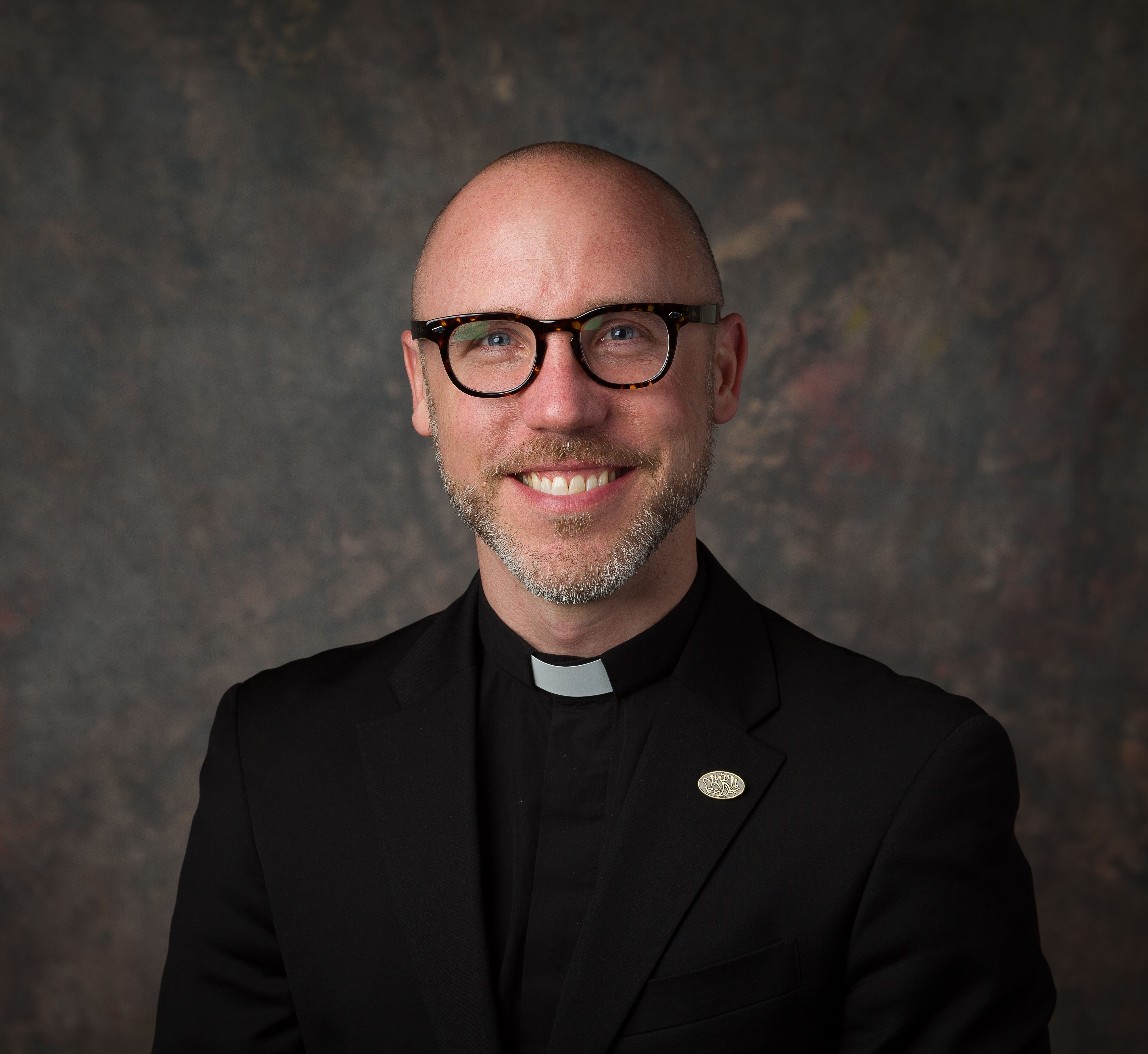 Man wearing glasses and a clerical collar smiling in a professional headshot against a dark background
