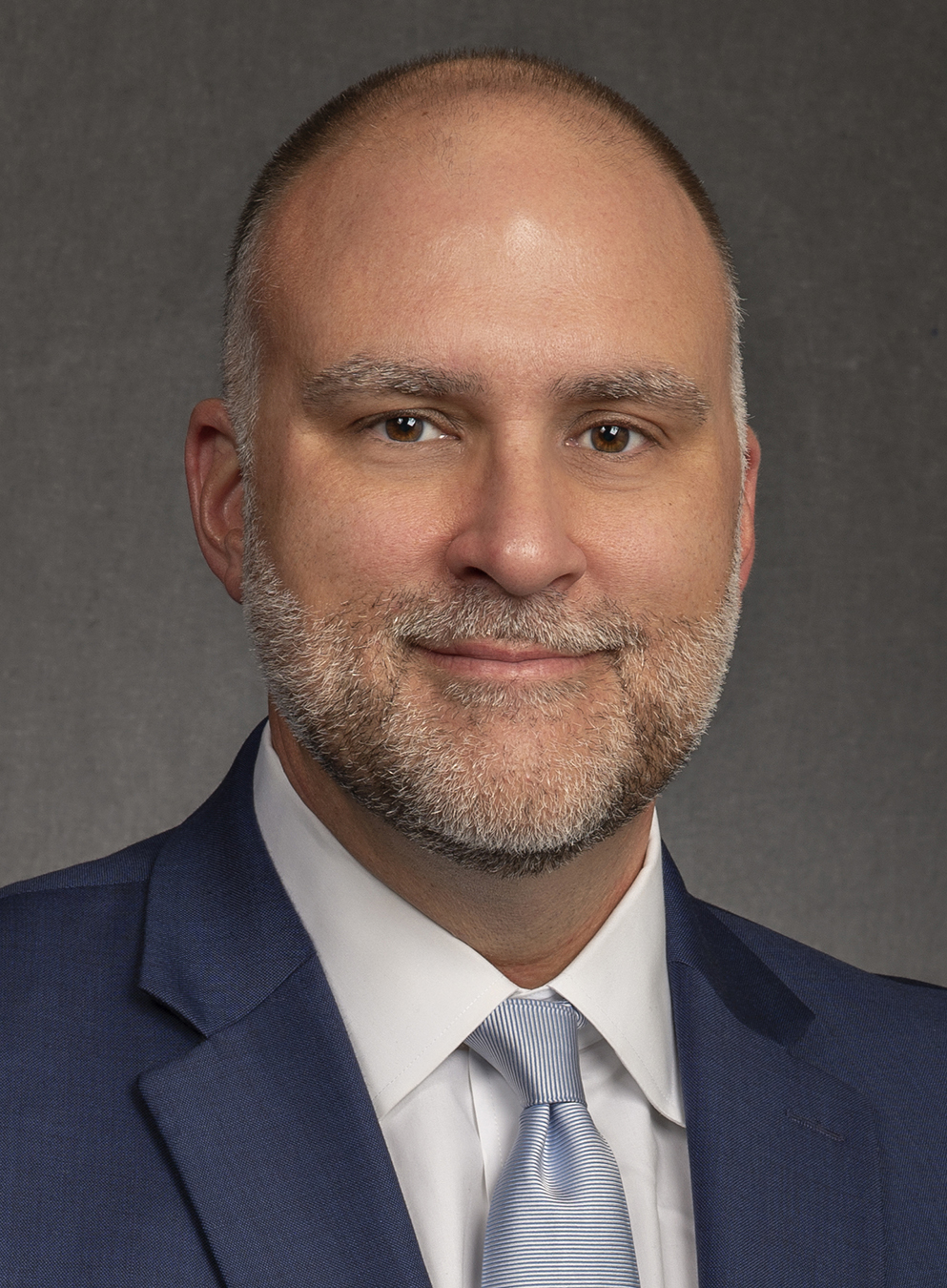 Man with a shaved head and short beard wearing a suit and tie, against a neutral background.