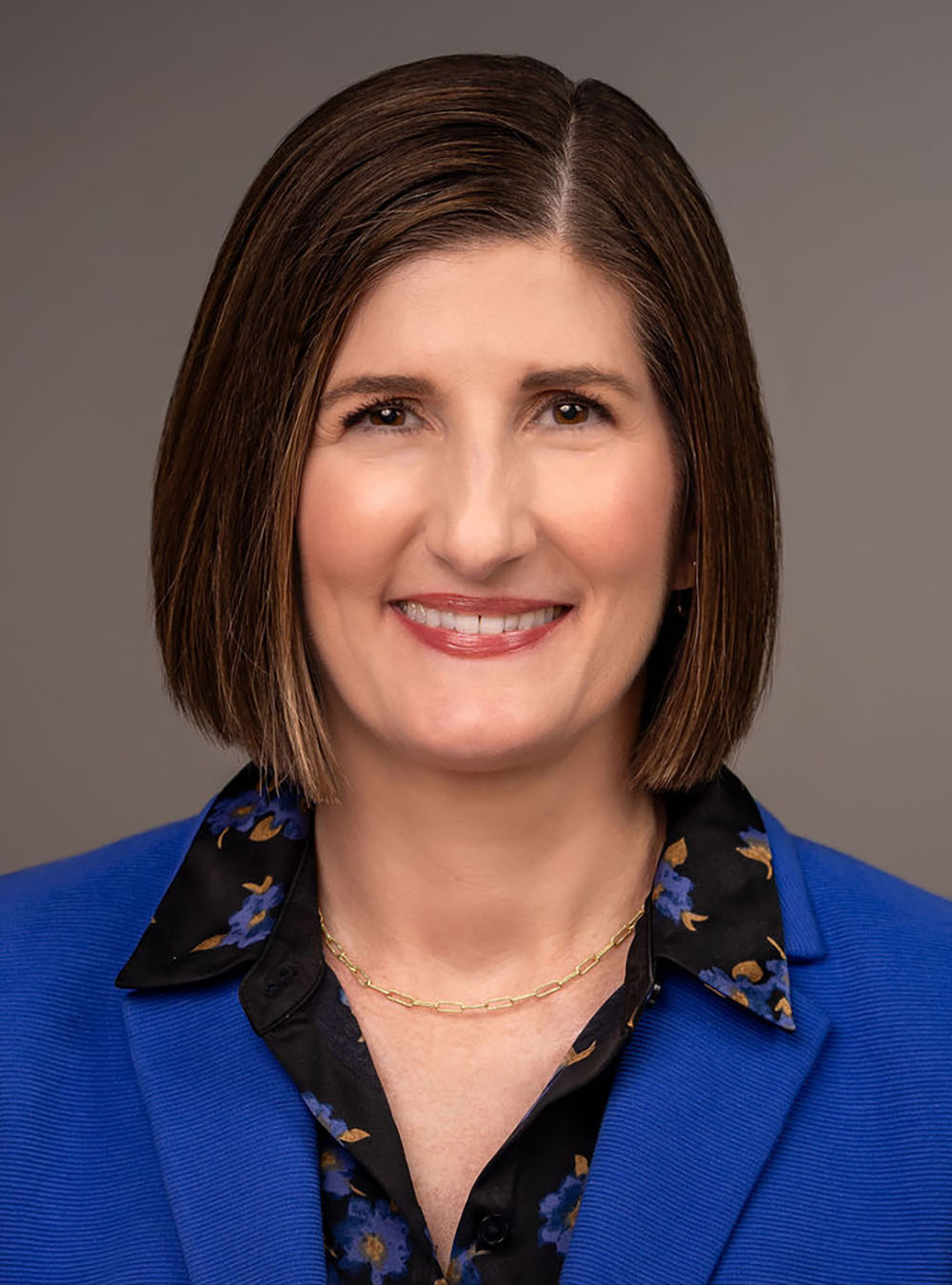 Woman with a short brown bob wearing a blue blazer, smiling against a neutral background.