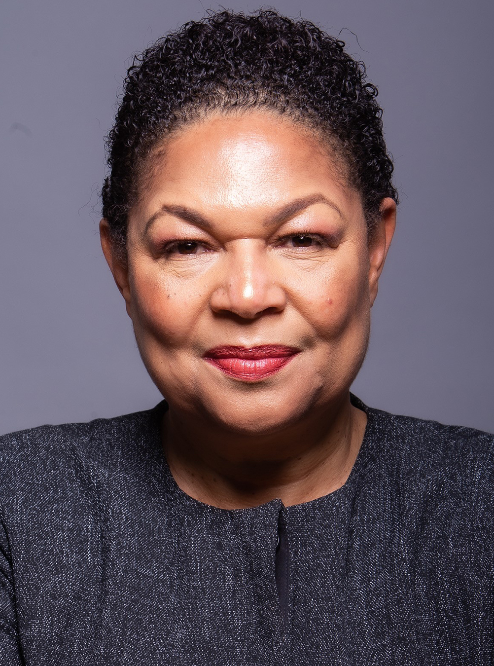 Woman with short curly hair wearing a dark top, looking directly at the camera against a neutral background.