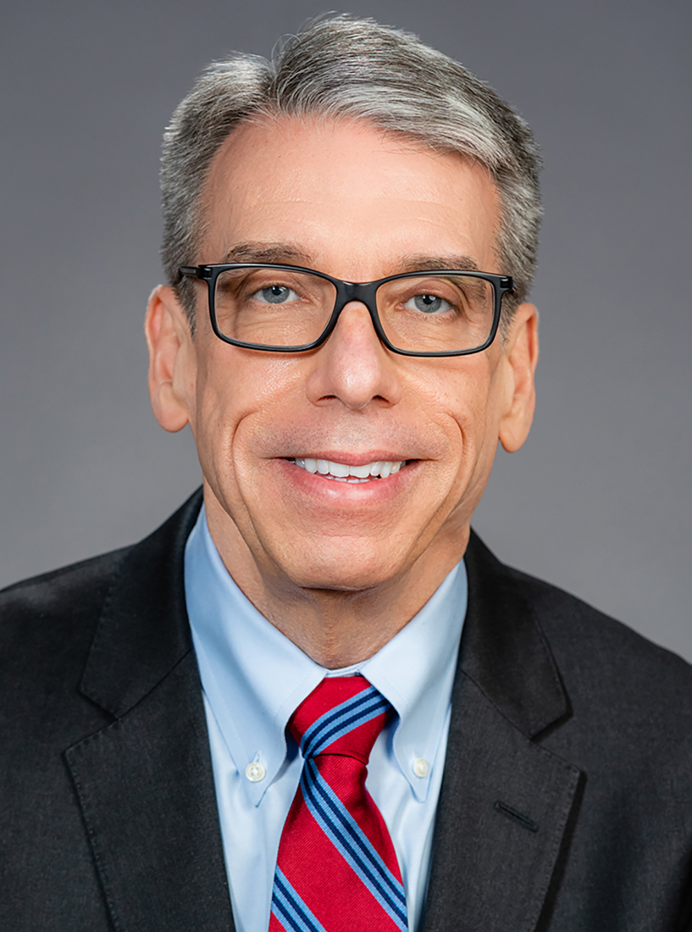 Smiling man with glasses in a suit and red striped tie against a plain background.