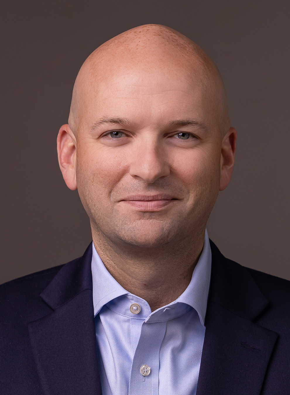 Man in a suit with a shaved head against a neutral background.