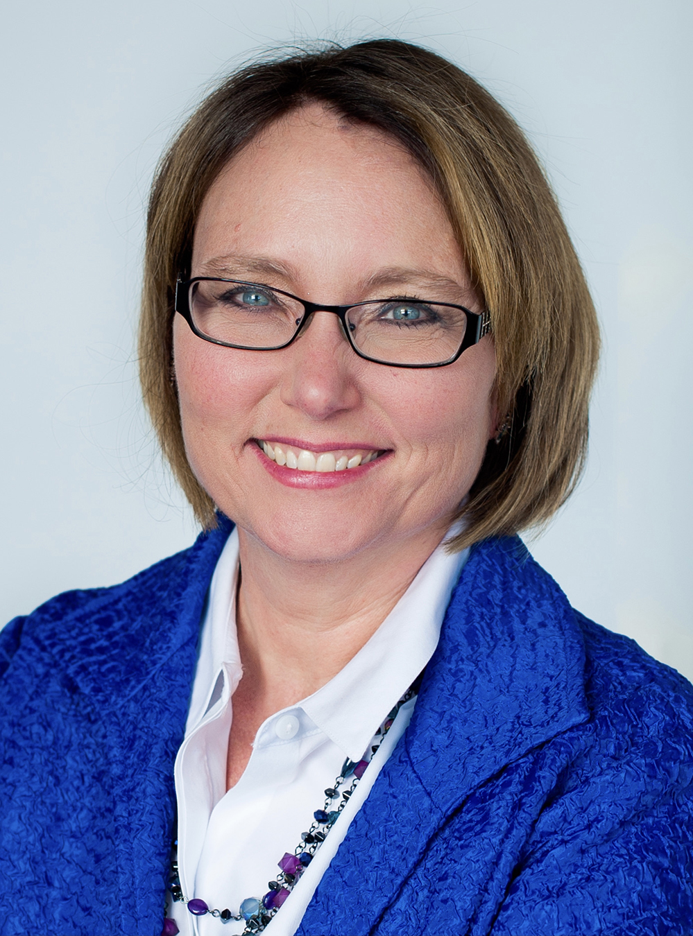 Woman with short light-brown hair and glasses, wearing a blue blazer and white blouse, smiling at the camera.
